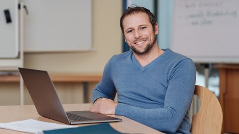 Lehrer sitzt mit Laptop am Pult.