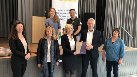 Staatssekretär Lösel mit Beteiligten der Diskussion vor einem Pult und einer Bühne, alle lächeln in die Kamera.