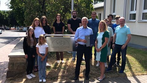 Staatssekretär Lösel besucht Lerncamp der Geschwister-Scholl-Schule Alsfeld. Er steht gemeinsam mit Schülerinnen und Schülern sowie Lehrerinnen und Lehrern vor dem Schild der Schule.