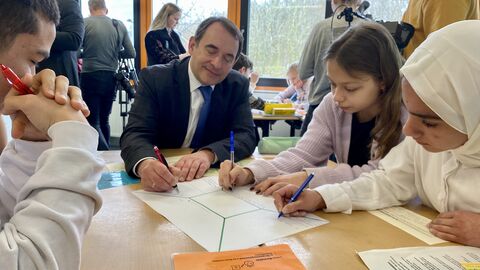 Kultusminister Lorz sitzt gemeinsam mit Schülerinnen und Schülern um einen Tisch. Alle haben Stifte in der Hand und schreiben auf ein blatt Papier.