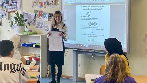 Eine Lehrerin steht am Whiteboard vor der Klasse und hält einen Zettel in der Hand. Die Schülerinnen und Schüler schauen aufmerksam zu.