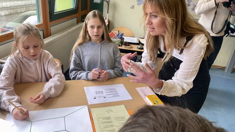 Die Lehrerin steht bei einer Gruppe Schülerinnen am Tisch und erklärt eine Aufgabe. Vor den Schülerinnen liegt ein Blatt Papier und eine Aufgabenstellung.