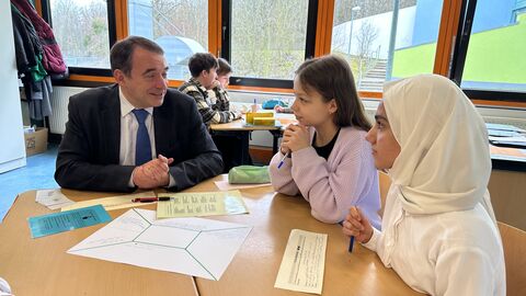 Kultusminister Lorz unterhält sich am Tisch sitzend mit zwei Schülerinnen, vor Ihnen liegen Aufgaben und Schreibmaterial. Im Hintergrund weitere Schüler an Gruppentischen.