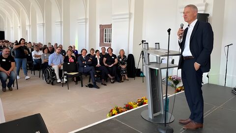 Staatssekretär Lösel auf einer Bühne vor Publikum. Er hält ein Mikrofon in der Hand und spricht.