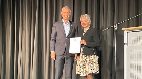 Staatssekretär Lösel steht neben Dr. Rosemarie zur Heiden auf einer Bühne neben einem Rednerpult. Im Hintergrund ein schwarzer, geschlossener Vorhang.