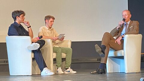 bildungsminister Schwarz sitzt in einem Sessel auf einer Bühne und spricht in ein Mikrofon. Ihm gegenüber sitzen zwei Schüler mit Moderationskarten.