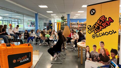 Blick in eine Schulklasse, die im Kreis sitzt. Im Vordergrund ein Aufsteller von "BÜCHERALARM @ school" sowie ein oranger Koffer mit der Aufschrift "Bücheralarm".