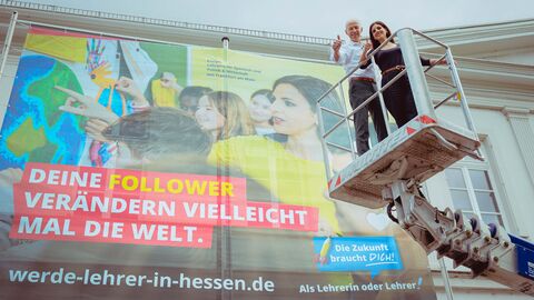 Von rechts auf einem Kran steht Kultusminister Schwarz gemeinsam mit einer Dame, hintendran an der Wand ist ein Plakat zu sehen mit der Aufschrift: "Deine Follower verändern vielleicht mal die Welt." der Initiative „Die Zukunft braucht Dich! Als Lehrerin oder Lehrer“.