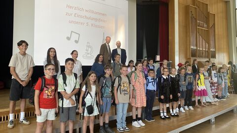 Bildungsminister Armin Schwarz steht gemeinsam mit Schülerinnen und Schülern auf einer Bühne. Auf einer Leinwand im Hintergrund steht: "Herzlich Willkommen zur Einweihungsfeier unserer 5. Klassen:"