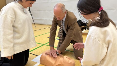 Ein Mann drückt auf den Brustkorb einer Puppe, um diese Wiederzubeleben, Zwei Schülerinnen schauen dabei zu.