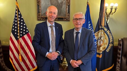 Bildungsminister Schwarz und Tony Evers stehen in einem Raum vor unter anderem der US-amerikanischen Flagge.