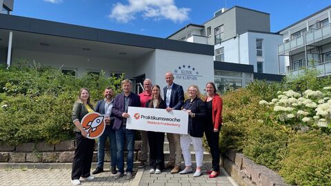 Mehrere Personen stehen vor Klinik und halten ein Schild in Kamera.