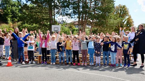 Eine große Gruppe von Schülerinnen und Schülern steht eng beieinander im Freien. Sie singen gemeinsam und haben dabei die Arme nach oben gestreckt.