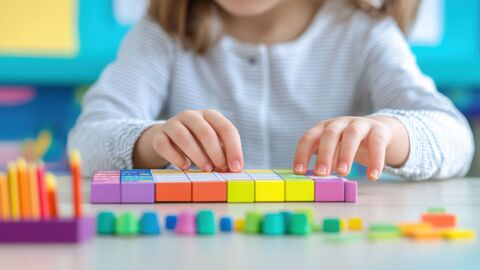 Ein Kind sitzt am Tisch und ordnet mit beiden Händen bunte Recenwürfel an.