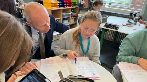 Ein Mann sitzt in einem Klassenraum mit einer Schülerin an einem Tisch und schaut ihr zu, wie sie etwas aufschreibt.