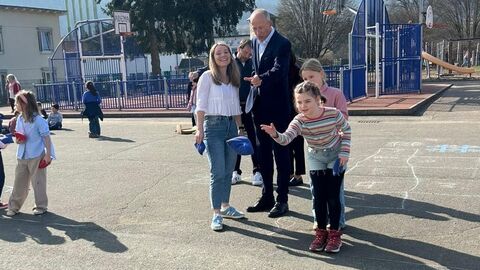 Minister Schwarz steht auf dem Pausenhof und unterhält sich mit einer Lehrerein. Um sie herum spielen Kinder. 