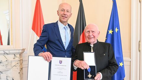Zwei Männer bei einer feierlichen Ordensverleihung vor Landes- und Bundesflaggen; einer hält die Urkunde, der andere präsentiert das Verdienstkreuz.