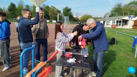 Erwachsene und Kinder stehen rund um einen Tisch an einem Hartplatz und halten Materialien für den Ballon in der Hand.