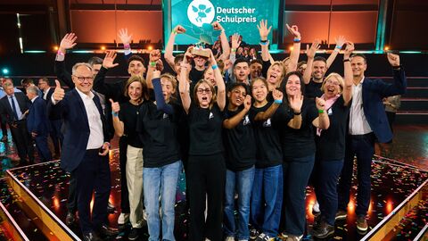Eine Gruppe Menschen, vor allem Schülerinnen und Schüler reißt die Arme hoch, dahinter ist das Logo des Deutschen Schulpreises zu erkennen